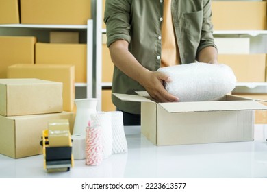 Young Small Business Entrepreneur Man, Online Store Owner With Shelves Of Boxes In The Background Packing Product In Mailing Box At Workplace For Shipping To Customer. Selling Product Online. 