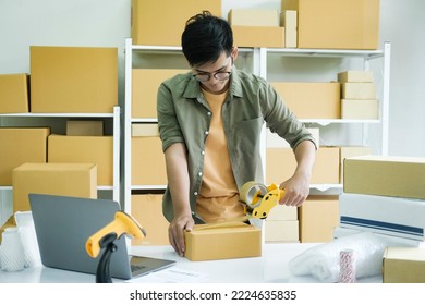 Young Small Business Entrepreneur Man, Online Store Owner With Shelves Of Boxes In The Background Packing Product In Mailing Box Sealing Box With Tape At Workplace For Shipping To Customer.