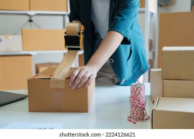 Young Small Business Entrepreneur Woman, Online Store Owner With Shelves Of Boxes In The Background Packing Product In Mailing Box Sealing Box With Tape At Workplace For Shipping To Customer.