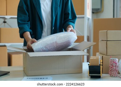 Young Small Business Entrepreneur Woman, Online Store Owner With Shelves Of Boxes In The Background Packing Product In Mailing Box At Workplace For Shipping To Customer. Selling Product Online.