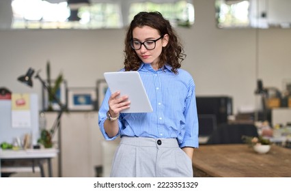 Young smart busy professional business woman executive, female company worker or manager holding digital tablet using pad technology device working standing in modern corporate office.