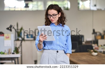 Young smart busy professional business woman executive, female company worker or manager holding digital tablet using pad technology device working standing in modern corporate office.