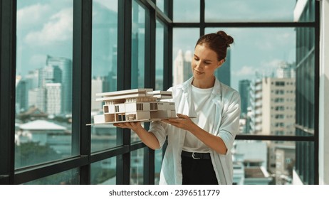 Young smart caucasian beautiful architect engineer raised house model while inspect carefully surrounded by city skyscraper view. Civil engineering, architectural studio. project planing. Tracery