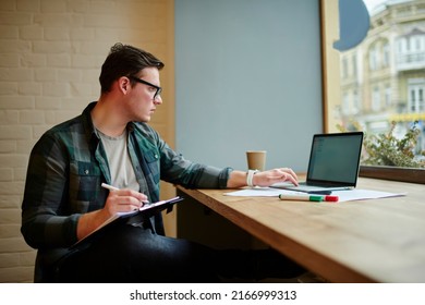 Young smart freelancer in glasses working with documents and laptop computer at desk. Male student learning online with netbook
