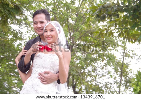 A young smart groom in black suit hold a bride in beautiful white dress with two red hearts in a park