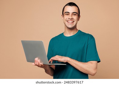 Young smart IT middle eastern man wear blue t-shirt casual clothes hold use work on laptop pc computer look camera isolated on plain pastel light beige background studio portrait. Lifestyle concept