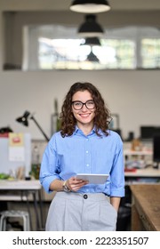 Young smart smiling professional business woman, happy confident 30s female company worker sales manager holding digital tablet standing in modern office working, looking at camera, vertical portrait.