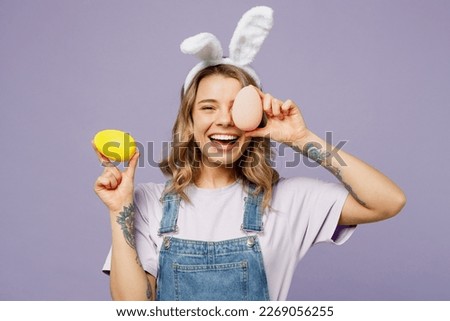 Young smiing cheerful fun woman wearing casual clothes bunny rabbit ears cover eye with colorful eggs isolated on plain pastel light purple background studio portrait. Lifestyle Happy Easter concept