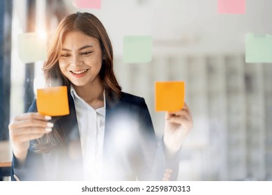 Young smiley attractive, businesswoman using sticky notes in glass wall to writing strategy business plan to development grow to success - Powered by Shutterstock