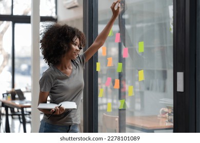 Young smiley attractive, businesswoman using sticky notes on a glass wall to write a strategic business plan to develop and grow to success. - Shutterstock ID 2278101089