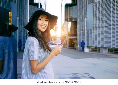 Young smiley well-dressed Asian business woman use digital tablet near skyscrapers. Flare light