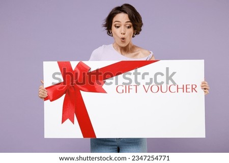 Young smilg happy surprised shocked amazed woman 20s with bob haircut wearing white t-shirt hold big gift certificate coupon voucher card for store isolated on pastel purple background studio portrait