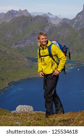 Young Smiling Active Man With Backpack Hiking On Lofoten Islands In Norway On Sunny Day High Above Fjord