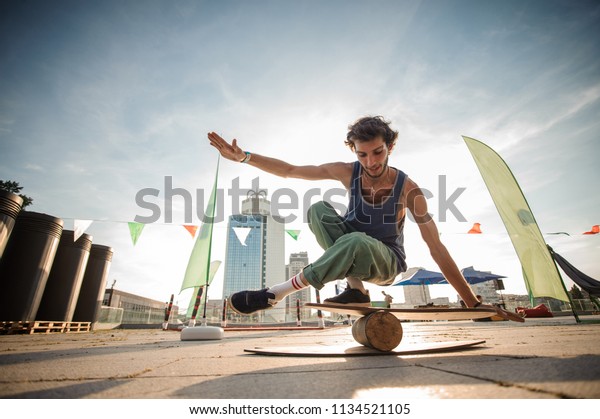 Young smiling\
active man keeping balance on the wooden board against the\
background of city buildings on summer\
day