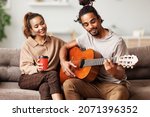 Young smiling african american man musician playing instumental music on guitar for happy girlfriend while sitting together on sofa and enjoying weekend at home, couple play on musicial instrument