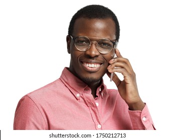 Young smiling african american man with round glasses on, pressing phone to ear while having conversation, isolated on white background 