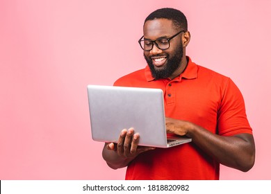 Young smiling african american man standing and using laptop computer isolated over pink background.