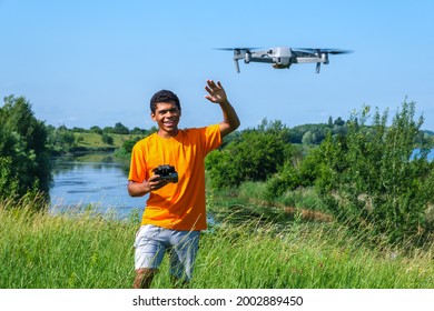 Young smiling African American man operating drone with controller in hands on the meadow near the river. 