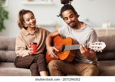 Young smiling african american man musician playing instumental music on guitar for happy girlfriend while sitting together on sofa and enjoying weekend at home, couple play on musicial instrument - Powered by Shutterstock