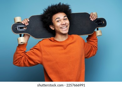 Young smiling African American man holding longboard isolated on blue background. Portrait of attractive happy skater looking at camera. Street culture, active lifestyle concept