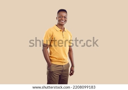 Young smiling African American man millennial with short hair posing with one hand in pocket pants and confidently looking at camera dressed in casual clothes stands on beige background. Copy space