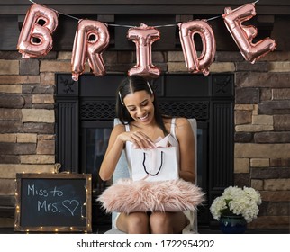 Young smiling African American woman opening a bridal shower present with pink bride balloons and brick fireplace in background
