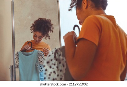 Young Smiling African American Woman Trying On Clothes In Front Of Mirror At Home, Happy Black Girl Holding Blue Summer Dress On Hanger, Deciding What To Wear For Walk Outside. Selective Focus