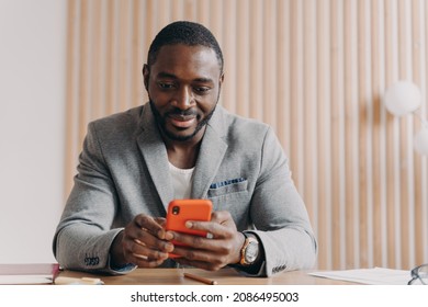 Young Smiling African Male Entrepreneur Sitting At Office Desk And Using Mobile Phone At Workplace, Handsome Successful Afro American Businessman Reading Online News Or Sending Email On Smartphone