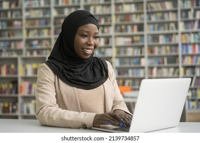 Young Smiling African Student Wearing Hijab Using Laptop Computer Studying Sitting In Modern Library, Education Concept. Beautiful Happy Nigerian Woman Typing On Keyboard Working Online At Workplace