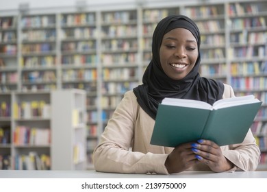 Young Smiling African Student Wearing Black Hijab Studying, Learning Literature Sitting In Modern Library, Education Concept. Beautiful Happy Muslim Woman Reading Book At Workplace 