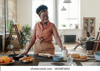 Young Smiling African Woman In Casualwear Standing By Electric Stove And Taking Bottle Of Milk While Cooking Dinner For Her Family In The Kitchen