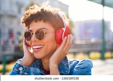 Young smiling african woman listening music with headphones at sunset. Brazilian girl listening songs via wireless headphones. Closeup face of teen wearing sunglasses and keeps the rhythm of the song.