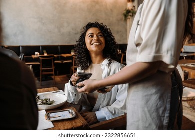 Young smiling african woman using smartphone while paying restaurant bill with contactless payment during dinner party with friends - Shutterstock ID 2322856651