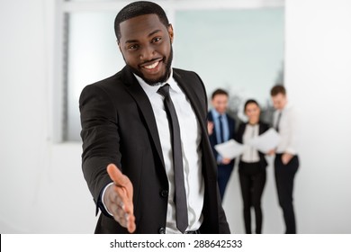 Young Smiling African-american Businessman Looking At Camera And Reaching Out His Hand. Business People Communicating In The Background