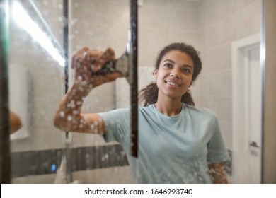 Young Smiling Afro American Woman In Uniform Cleaning Shower Door With Squeegee And Spray Detergent. Cleaning Service Concept. Housework