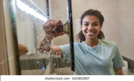 Young Smiling Afro American Woman In Uniform Cleaning Shower Door With Squeegee And Spray Detergent. Cleaning Service Concept. Housework