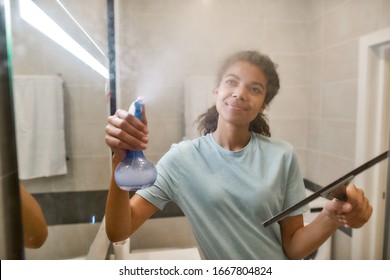 Young Smiling Afro American Woman In Uniform Cleaning Shower Door With Squeegee And Spray Detergent. Cleaning Service Concept. Housework