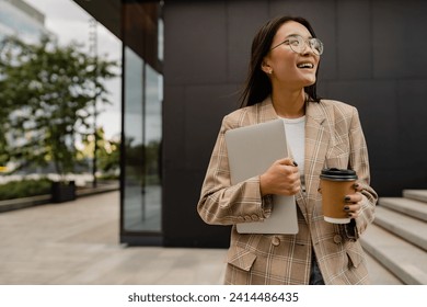 young smiling asian attractive woman walking outside in street doing business, education, student working, summer casual style, holding laptop and drinking coffee, wearing glasses, beige jacket  - Powered by Shutterstock