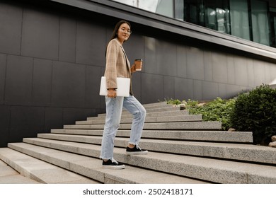 young smiling asian attractive woman walking outside in street doing business, education, student working, summer casual style, holding laptop and drinking coffee, wearing glasses, beige jacket  - Powered by Shutterstock