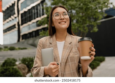 young smiling asian attractive woman walking outside in street doing business, education, student working, summer casual style, holding laptop and drinking coffee, wearing glasses, beige jacket - Powered by Shutterstock