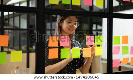 Young smiling asian business woman mentor leader write tasks creative ideas on sticky post it notes on glass wall, vietnamese female coach planning project corporate management on sticker scrum board