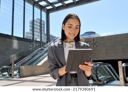 Young smiling Asian business woman entrepreneur wearing suit holding digital tablet standing in city metro using wireless internet technology on pad computer, working online in subway.