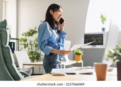 Young smiling Asian businesswoman working at modern office, standing by workdesk with laptop, cheerful lady holding paper documents making phone call, having cellular conversation, copy space - Powered by Shutterstock