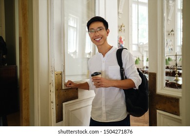 Young smiling asian male student in eyeglasses with backpack and coffee to go opening library door in university