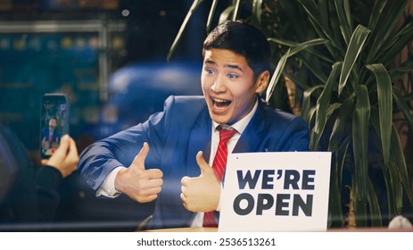 Young smiling Asian man in blue suit taking photo with we are open sign and thumbs-up, showing excitement about opening new venture. Concept of new business, startup, achievement, progress - Powered by Shutterstock