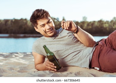 Young Smiling Asian Man Lying On Sandy Beach With Bottle Of Beer And Looking At Camera