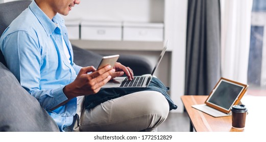 Young Smiling Asian Man Relaxing Using Laptop Computer Working And Video Conference Meeting At Home.Young Creative Man Looking At Screen Typing Message With Smartphone.work From Home Concept