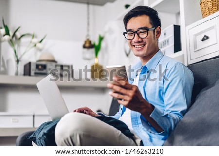 Young smiling asian man relaxing using laptop computer working and video conference meeting at home.Young creative man looking at screen typing message with smartphone.work from home concept