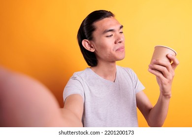 Young Smiling Asian Man Talking On Video Call, Enjoying Coffee First Person View. Asian Teenager Chatting On Videoconference, Holding Mobile Phone, Drinking Tea To Go Pov