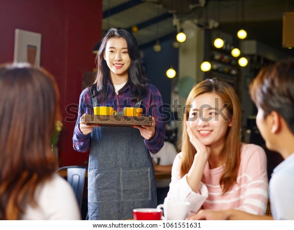young smiling asian waitress serving\
customers in coffee shop, focus on the background\
girl.